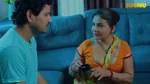 Two actors in a scene from a Jugnu web series, sitting on the floor near a blue sofa, a woman in an orange saree holding a mug